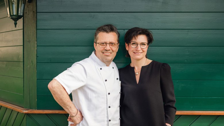 Silvia und Albin Hawel, © Niederösterreich Werbung/Daniela Führer Ein Mann in Kochjacke und eine Frau in schwarzem Kleid stehen vor einer grünen Holzwand.