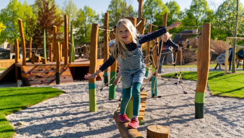 Starkl Erlebnisgarten, © starklfilm Ein Kind balanciert auf einem Holzstamm auf einem Spielplatz mit Klettergerüsten im Hintergrund.