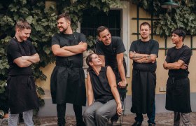 Tradition und Innovation und Teamgeist, © Niederösterreich Werbung/Daniela Führer Eine Gruppe von sechs Personen in schwarzer Arbeitskleidung steht und sitzt lächelnd vor einer mit Weinreben bewachsenen Wand.