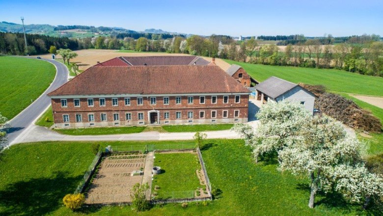 Moststraße von oben, © Cleanhill Studios Luftaufnahme eines Bauernhofs mit roten Ziegeldächern, umgeben von Feldern und blühenden Bäumen.