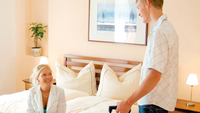Gestehof Hetzenberg, © Gestehof Hetzenberg Ein Mann und eine Frau in einem Hotelzimmer, die Frau sitzt auf dem Bett, der Mann steht mit einem Koffer.