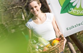 Rundum Gesund-Shop, © schwarz-koenig.at Frau mit Korb voller Gemüse vor einem Schild mit der Aufschrift 'gesund'.