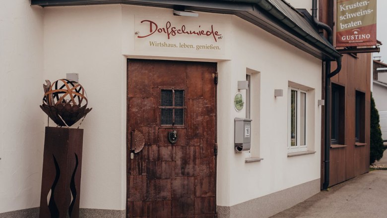 Eingang eines Wirtshauses namens Dorfschmiede mit rustikaler Tür und Dekoration.