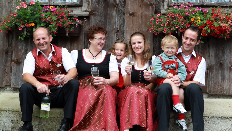 Eine Familie in traditioneller Kleidung sitzt vor einem Holzhaus mit Blumen und hält Weingläser.