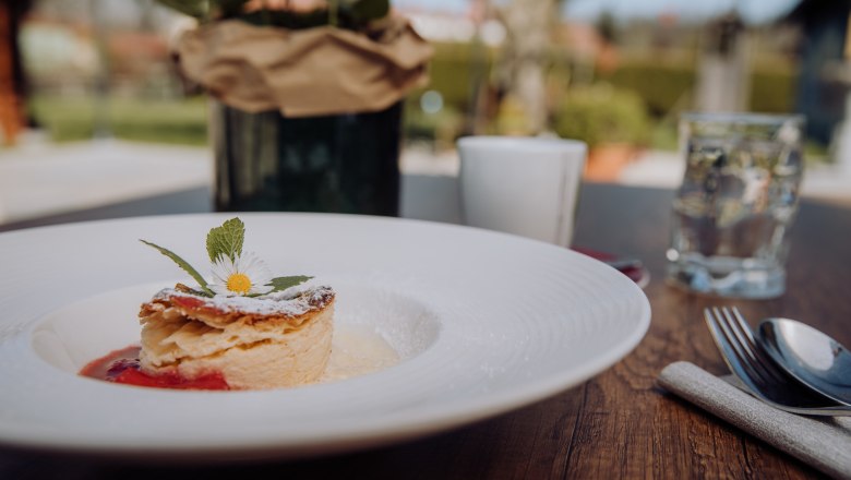 Ein Teller mit Milchrahmstrudel, garniert mit einem G&auml;nsebl&uuml;mchen, auf einem Holztisch im Freien.