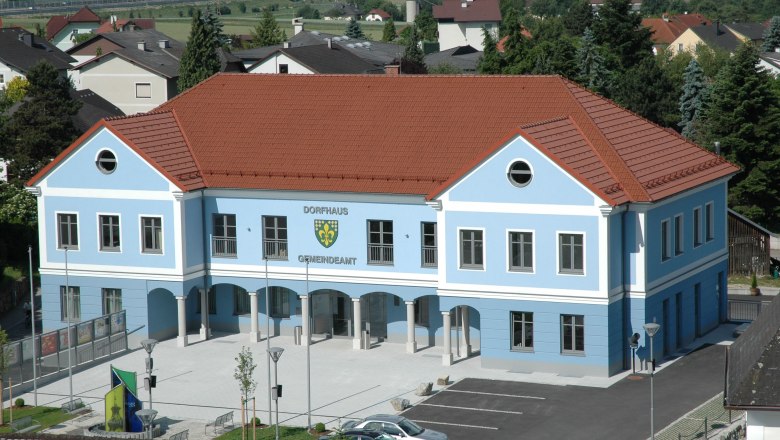 Blaues Gebäude mit rotem Dach, beschriftet als Dorfhaus Gemeindeamt, umgeben von Häusern und Bäumen.