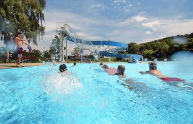 Kinder schwimmen im Freibad Neuhofen an der Ybbs mit einer großen Wasserrutsche im Hintergrund.