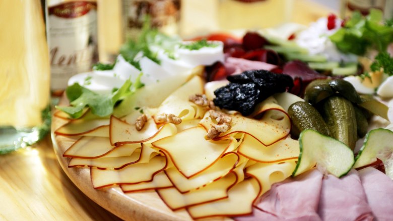Ein Jausenbrett mit K&auml;se, Schinken, Gurken, N&uuml;ssen und Salat auf einem Holztisch.