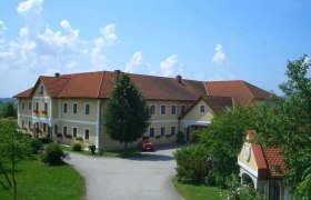 Großer Bauernhof mit gelben Wänden und roten Dächern, umgeben von Bäumen und einer Straße.