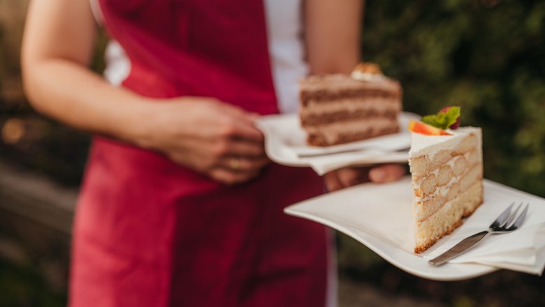 Person in roter Schürze hält zwei Teller mit Tortenstücken.