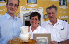 Drei Personen vor einem gelben Gasthaus mit einem Tablett und einem alten Foto.