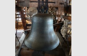 Gro&szlig;e Glocke in einem h&ouml;lzernen Glockenstuhl, umgeben von Steinmauern.
