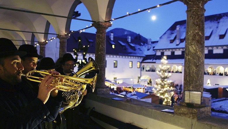 Musiker spielen Trompeten auf einem Balkon mit Blick auf einen Weihnachtsmarkt bei Nacht.