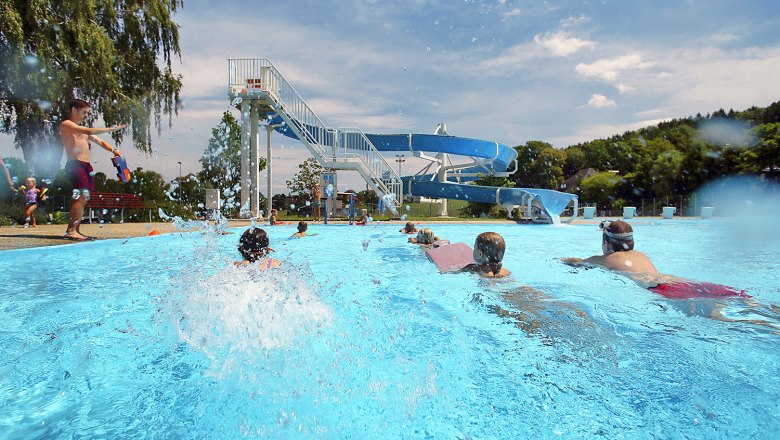 Kinder schwimmen im Freibad Neuhofen an der Ybbs mit einer großen Wasserrutsche im Hintergrund.