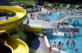 Ein Freibad mit gelber Wasserrutsche, Schwimmern im Becken und Menschen auf der Liegewiese.