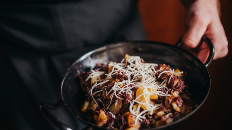 Eine Person h&auml;lt eine Pfanne mit Blunzengr&ouml;stl, garniert mit geriebenem K&auml;se.