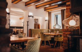 Gem&uuml;tliches Restaurant mit Holzbalken, gr&uuml;nen Polsterst&uuml;hlen und dekorativen Lampen.