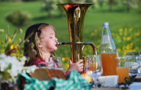 Ein kleines Mädchen spielt eine Tuba auf einer Wiese, umgeben von Getränken und Blumen.