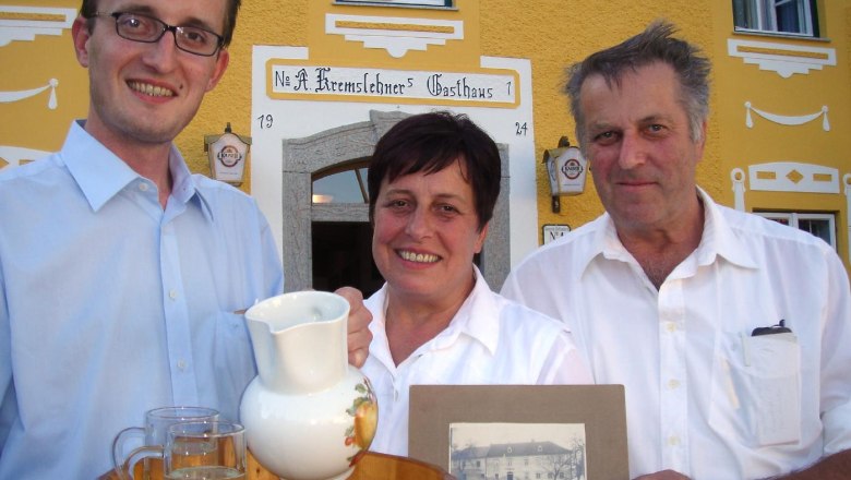 Drei Personen vor einem gelben Gasthaus mit einem Tablett und einem alten Foto.