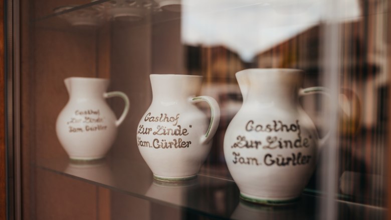 Drei Kr&uuml;ge mit Aufschrift "Gasthof Zur Linde Fam. G&uuml;rtler" in einer Vitrine.