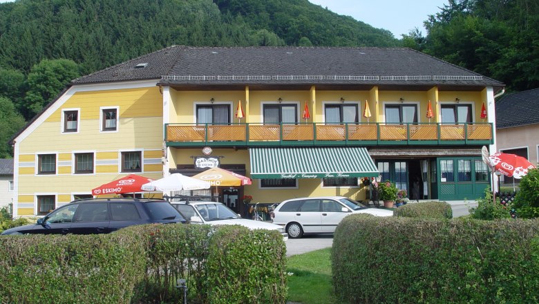 Gelbes Gasthaus mit Balkon und Sonnenschirmen vor einem bewaldeten H&uuml;gel.