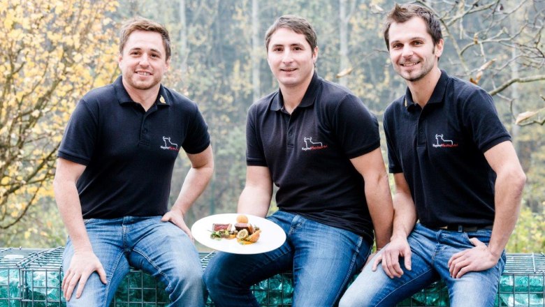 Drei Männer in schwarzen Poloshirts sitzen draußen und halten einen Teller mit Essen.