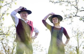 Zwei Personen in traditioneller Kleidung mit H&uuml;ten stehen in einem bl&uuml;henden Obstgarten.