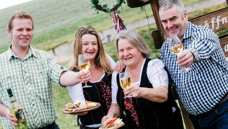 Vier Personen sto&szlig;en mit Weingl&auml;sern an, im Freien vor einem Schild.
