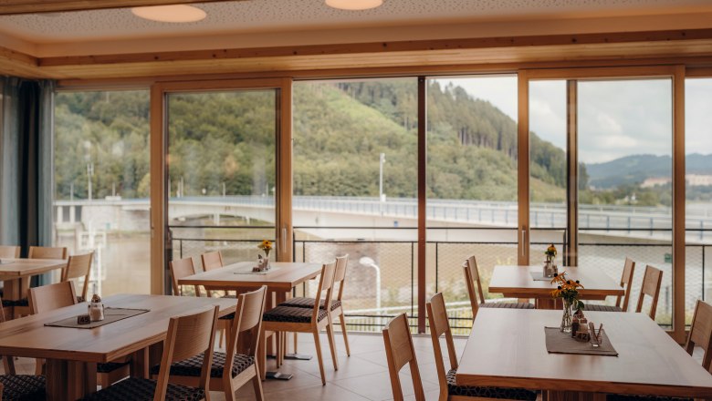 Innenansicht eines Gasthofes mit gro&szlig;en Fenstern und Blick auf eine Br&uuml;cke und bewaldete H&uuml;gel.