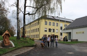 Gruppe von Menschen vor einem gelben Schulgebäude mit Solarpanelen auf dem Dach.