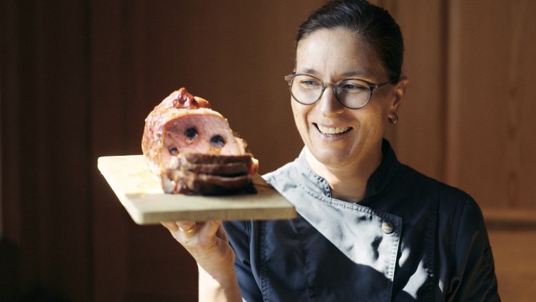 Eine Frau in Kochkleidung h&auml;lt ein Holzbrett mit einem aufgeschnittenen Fleischgericht.