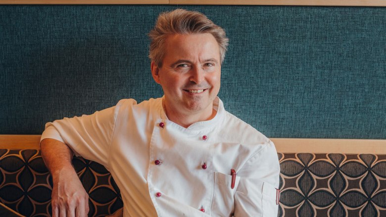 Ein l&auml;chelnder Mann in wei&szlig;er Kochjacke sitzt auf einer gepolsterten Bank vor einer gemusterten Wand.