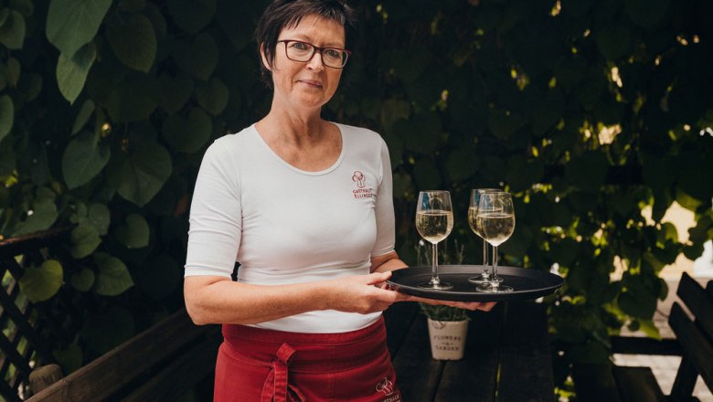 Frau mit Brille und Tablett mit zwei Weingläsern im Freien.