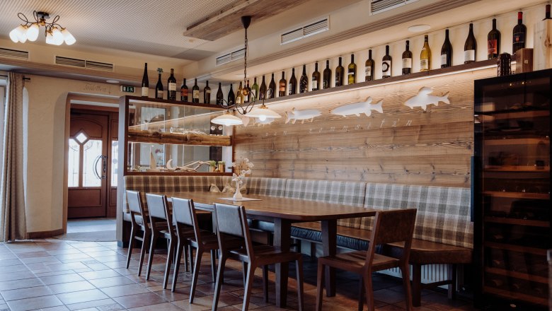 Gem&uuml;tlicher Essbereich mit Holztisch, St&uuml;hlen und Bank, dekoriert mit Weinflaschen und Fischmotiven an der Wand.