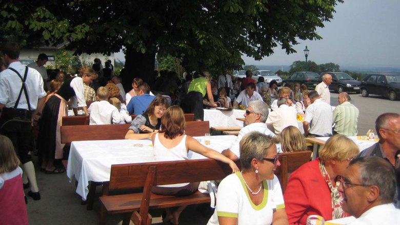 Menschen sitzen an langen Tischen im Freien unter einem Baum.