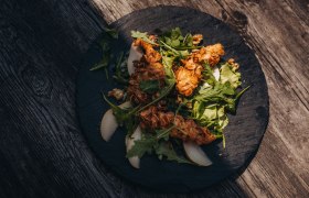 H&uuml;hnerfilet mit Mandelkruste auf Salat mit Birnenscheiben auf einem schwarzen Teller.