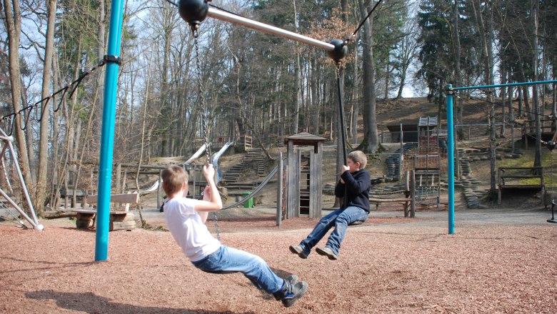 Zwei Kinder schaukeln auf einem Seilspielgerät auf einem Spielplatz im Wald.