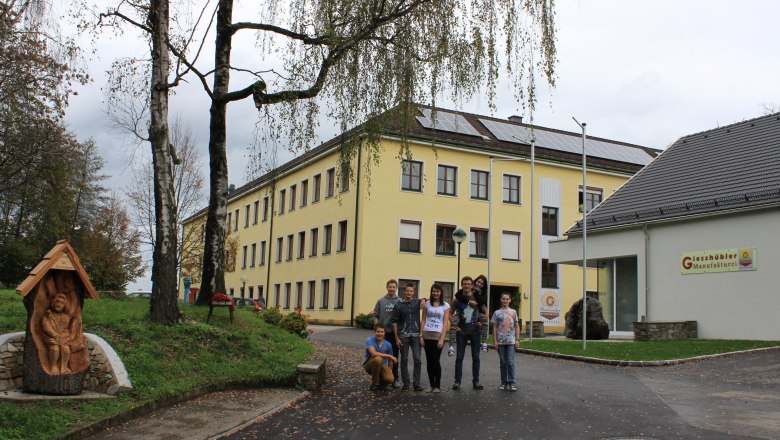Gruppe von Menschen vor einem gelben Schulgebäude mit Solarpanelen auf dem Dach.