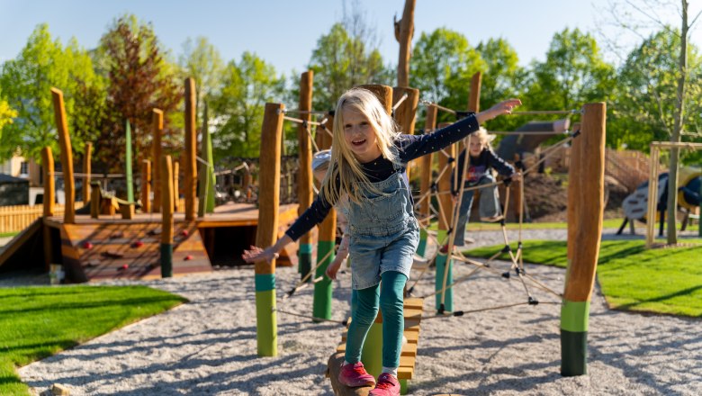 Ein Kind balanciert auf einem Holzstamm auf einem Spielplatz mit Kletterger&uuml;sten im Hintergrund.