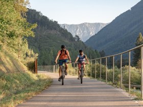 Zwei Radfahrer gleiten entspannt entlang des Ybbstalradwegs, umgeben von der majest&auml;tischen Kulisse der Ybbstaler Alpen. Die sanften H&uuml;gel und das klare, blaue Himmel schaffen eine einladende Atmosph&auml;re f&uuml;r Abenteuerlustige und Naturliebhaber. Hier wird der Sommer in den Bergen zum unvergesslichen Erlebnis.