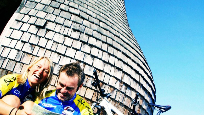 Zwei Radfahrer sitzen vor einer großen birnenförmigen Holzkonstruktion und lachen.