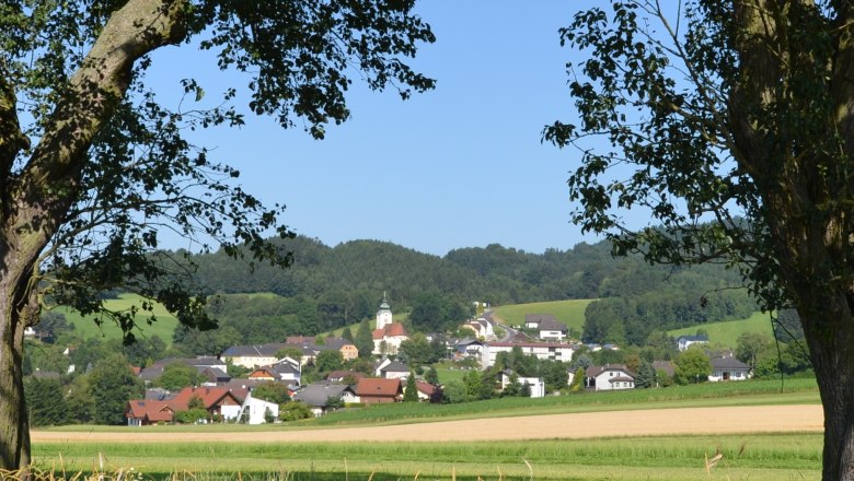 Landschaft mit Dorf und Kirche im Hintergrund, umrahmt von Bäumen.